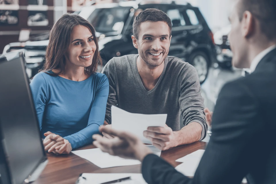Couple souriant assis face &agrave; un conseiller dans un showroom automobile, examinant des documents contractuels sur une table en bois